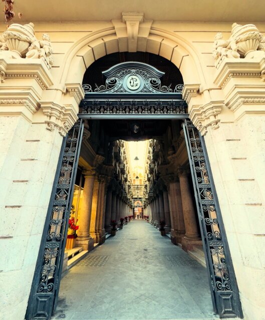 Pasaje comercial cubierto visto desde la puerta abierta, hierro forjado en filigrana sobre el dintel y profunda perspectiva de columnas hasta una salida lejana. Una figura mínima al fondo da la escala. Arquitectura decimonónica para gente que aún tenía tiempo de pasear bajo techo.