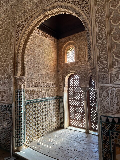Interior nazarí con arco de herradura abierto sobre una sala más pequeña, yesería tupida cubriendo cada palmo del muro con caligrafía y arabescos. Alicatado geométrico verde, blanco y azul al zócalo, celosía de madera al fondo dejando entrar la luz en cuadrícula. La habitación entera escribe el mismo poema repetido en cien variaciones, por si Dios no escuchó la primera vez.