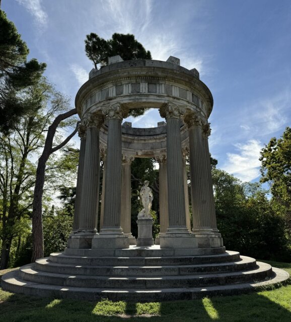 Templete circular de inspiración clásica sobre escalinata, columnas estriadas y entablamento sostenido por seis fustes. Una figura desnuda en mármol ocupa el centro, custodiada por la sombra del pino. Capricho aristocrático que el tiempo convirtió en plano de jardín público.