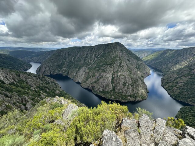 Un meandro fluvial cerrado abraza una mole de monte cubierta de bosque denso; el cielo cargado se cierra sobre los riscos. En primer plano, la roca y el matorral asoman al borde del precipicio. La curva del agua precede a quien la mira por miles de años.