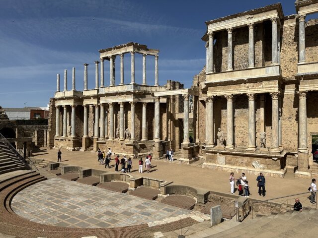 Teatro romano de escena alta con dos órdenes de columnas corintias en piedra clara, frontones partidos y nichos con estatuas. Pequeñas figuras de visitantes en la orchestra dan escala al monumento. Veinte siglos después, el público sigue siendo el de siempre: gente que llega tarde y se apoya contra la columna del fondo.
