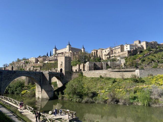 Vista panorámica de una ciudad amurallada coronando un cerro, alcázar cuadrado en lo alto con cuatro torres en las esquinas. En primer plano, un puente de piedra de dos arcos cruza un río verde flanqueado por una pasarela de madera con caminantes. Las ciudades sobre roca aprenden pronto que su mejor defensa es no ser cómodas.