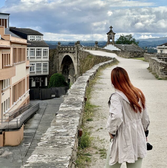 Mujer pelirroja con cazadora clara caminando por el adarve de una muralla romana, cámara colgando del hombro. Al fondo, un arco de entrada y una capillita con espadaña y cruz. La piedra cuadrada lleva veinte siglos sosteniendo el paseo de los aburridos del lunes por la mañana.