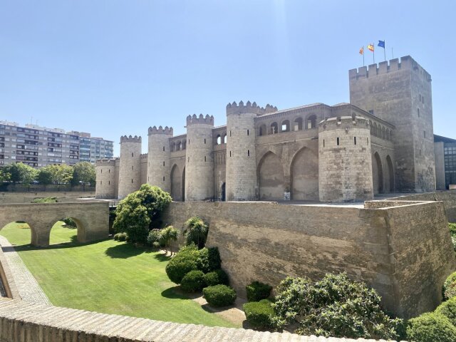 Palacio fortificado de muros amarillos con torres semicirculares almenadas y arquería ciega, una torre cuadrada más alta a la derecha con tres banderas en lo alto. Foso seco convertido en jardín, arcos de medio punto bajo el muro principal. Lo que fue residencia palaciega, prisión y cuartel hoy alberga otro tipo de poder: la piedra lleva mejor la rotación de funciones que sus inquilinos.