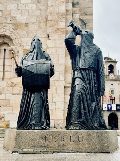 Las dos figuras del Merlú en bronce sobre un pedestal con la palabra MERLÚ grabada: encapuchadas, sin rostro, una sostiene un tambor a la cintura y un palillo, la otra alza una trompeta hacia el cielo. Detrás, un muro de catedral románica y, al fondo, un campanario con cigüeña. La procesión convertida en monumento queda muda.