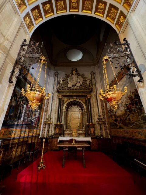 Interior de una capilla: arco artesonado en oro y rojo, retablo central de madera oscura con dos angelotes, dos arañas de bronce gigantes con campanas de vidrio rosa colgadas con cadenas larguísimas, y una alfombra roja saturada que cubre todo el suelo hasta el altar. Pinturas murales a los lados. Tanto oro alrededor para sostener, en realidad, un silencio.
