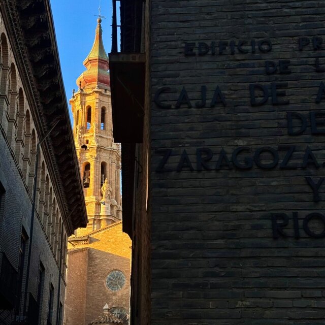 Callejón en sombra: a la derecha, una pared de ladrillo oscuro con letras en relieve que recortan EDIFICIO… CAJA DE A… DE ZARAGOZA Y RIO…; a la izquierda, asomando entre tejados, una torre barroca con tejas vidriadas verdes y naranjas iluminada por el sol bajo. Solo el campanario recibe luz; el banco, en sombra. La ciudad reparte el oro como cualquier otro presupuesto.