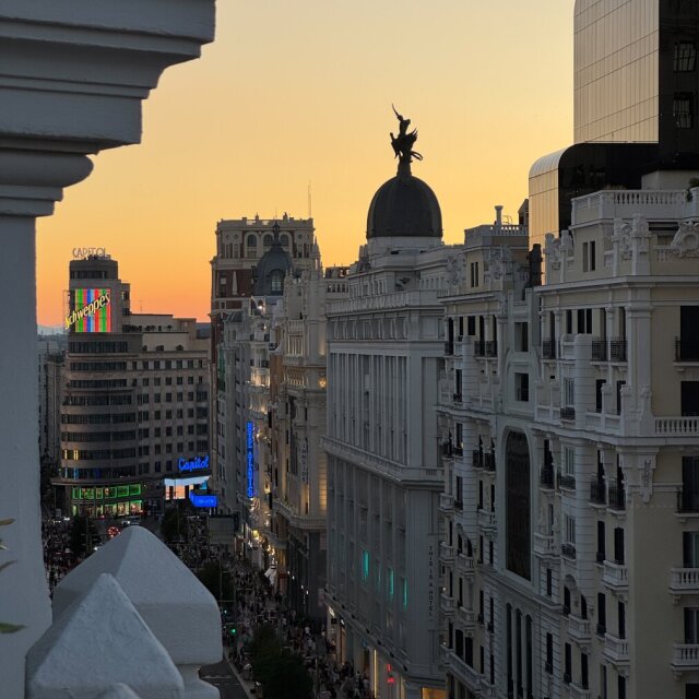 Atardecer sobre la Gran Vía visto desde un balcón blanco en primer plano: a la izquierda, el edificio Capitol con un letrero verde de Schweppes ya encendido; al centro, la cúpula negra del Metrópolis con la Victoria alada en silueta contra cielo melocotón; a la derecha, fachadas blancas con balcones. Dos siglos pidiendo permiso al mismo cielo y los dos lo consiguen.