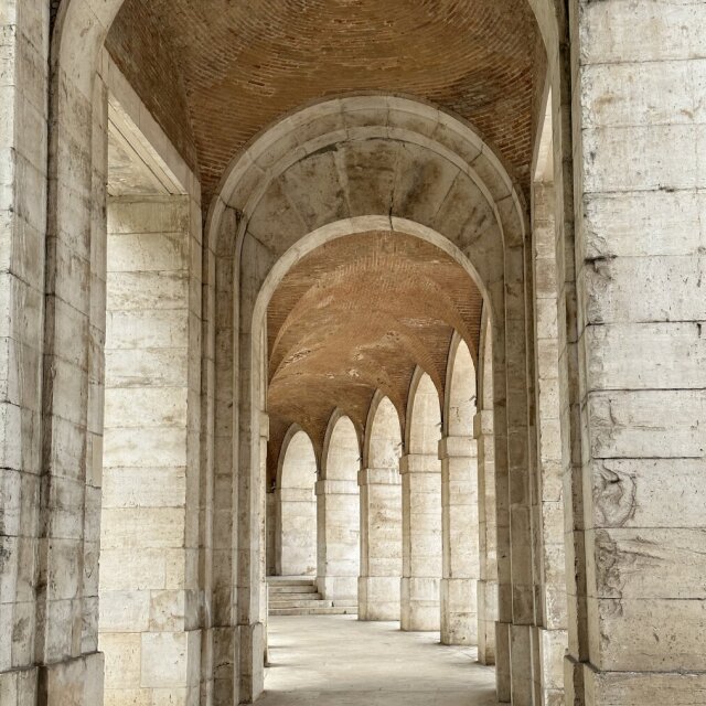 Galería abovedada con una sucesión de arcos de medio punto que se enfilan en perspectiva; los pilares de piedra clara sostienen una bóveda de ladrillo visto. La luz entra blanda por los lados y la profundidad se va estrechando arco tras arco. Cada vano es otro umbral hacia el siguiente.