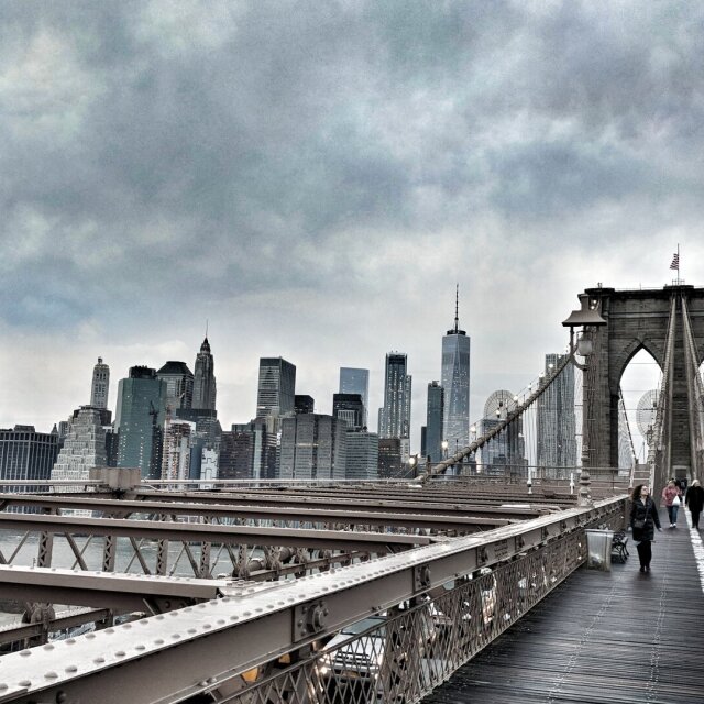 Pasarela peatonal del Puente de Brooklyn vista al frente: tablones gastados, cables verticales tensos hacia las almenas de piedra de la torre, una bandera diminuta en lo alto. Más allá, el skyline bajo un cielo de nubes pesadas y, a un lado, dos siluetas caminando hacia el otro lado. Cruzar es la forma más honesta de leer una ciudad.