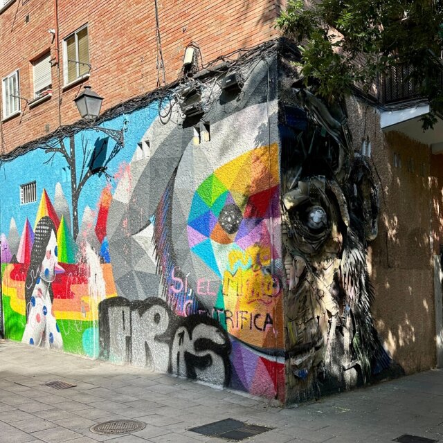 Esquina urbana cubierta por dos murales: a la izquierda, una composición poligonal de colores planos al estilo Okuda; a la derecha, una figura escultórica hecha enteramente de plásticos, neumáticos y metal recuperado, pegada al muro como cara de basurero. En medio, un graffiti negro tapa un cartel anterior. La calle se confiesa en la pared antes que en cualquier otra parte.