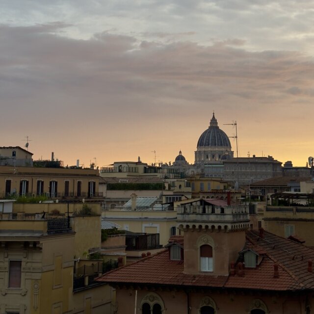 Tejados de Roma al atardecer: tejas curvas en hileras, terrazas con plantas, antenas viejas, mansardas y, al fondo, la cúpula de San Pedro silueteada en gris contra un cielo que pasa del lila al dorado. La ciudad parece detenerse a respirar. La cúpula funciona aquí como punto y aparte de la frase larga que es el horizonte.