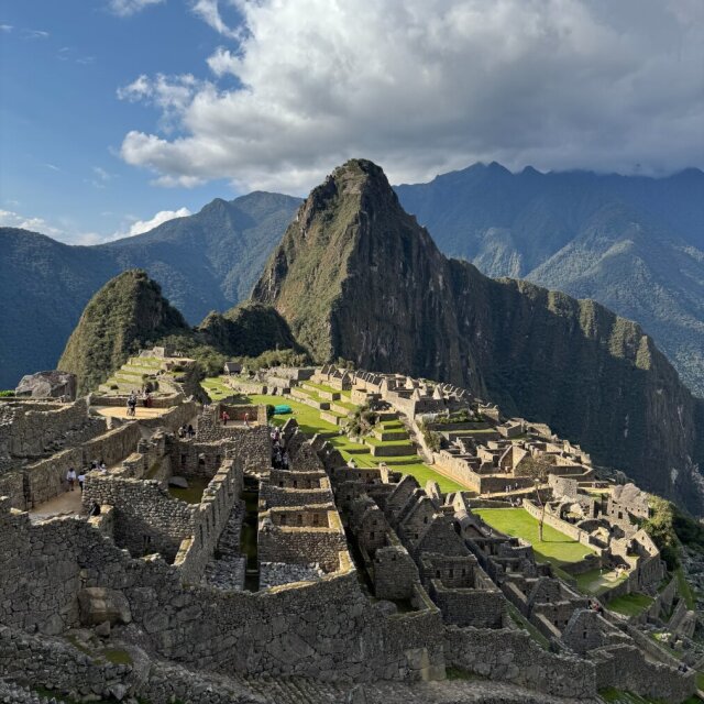 Las ruinas escalonadas de Machu Picchu en su silla de montaña: terrazas cortadas como peldaños, muros de granito sin mortero, calles internas mínimas. Detrás, el Huayna Picchu como cuerno verde y, más allá, cordilleras desvaneciéndose en niebla. Alguien decidió un día que el santuario quedaba mejor justo aquí, donde el aire ya cuesta.