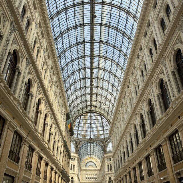 Interior de la Galleria Umberto I: bóveda inmensa de vidrio y hierro forjado dividida en cuadrículas regulares, dos plantas de fachadas eclécticas con balconadas y, en el fondo, una cúpula central que termina la perspectiva. Mucha gente diminuta abajo. El siglo XIX inventando catedrales sin santo, solo para que el comercio tuviera nave.