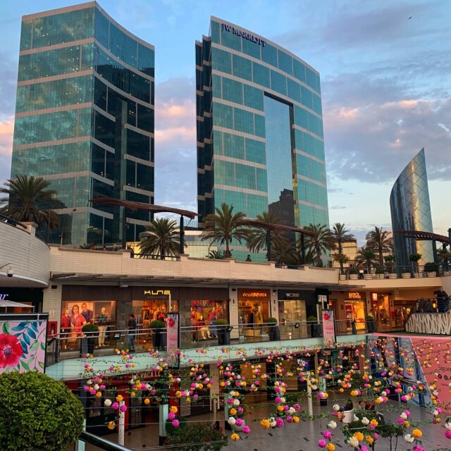 Dos torres del JW Marriott en cristal azul verdoso contra un cielo de atardecer en rosa y celeste; al lado, un edificio menor en cristal espejado y un voladizo blanco de centro comercial. Abajo, escaparates encendidos y un balcón decorado con guirnaldas de flores rosa, blancas y naranjas. El lujo intentando lavarse la cara con flores artificiales.