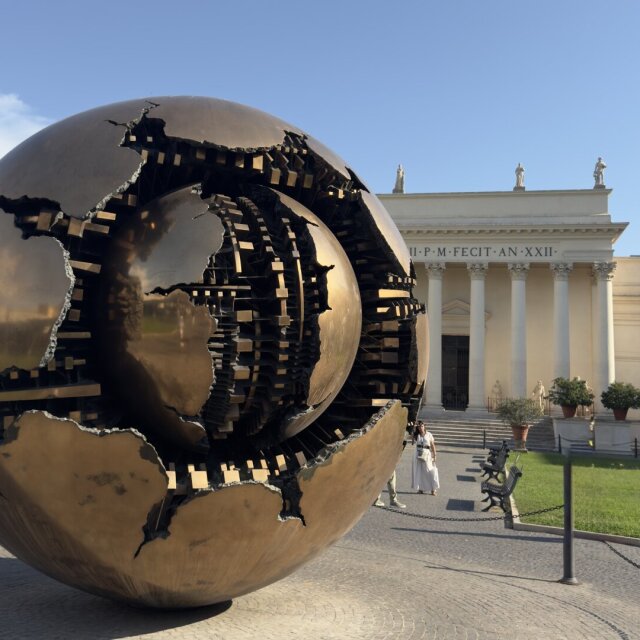 Sfera con sfera de Pomodoro en un patio del Vaticano: una esfera de bronce dorado abierta a grietas profundas, dejando ver dentro otra esfera más pequeña hecha de engranajes y láminas. Detrás, una fachada neoclásica con inscripción latina y, junto a la pieza, una pareja con vestido de novia posando. El mundo se rompe y dentro hay otro mundo idéntico, también roto.