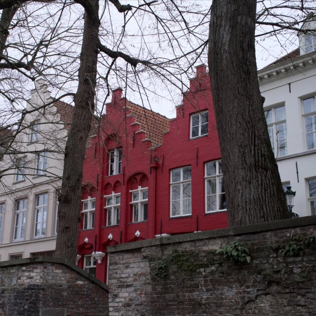 Una fachada roja con dos hastiales escalonados se asoma entre los troncos grises de dos árboles sin hojas; a la derecha, una casa blanca de molduras claras la acompaña en silencio. Al pie, un muro de ladrillo viejo cierra el jardín. El rojo es lo que más calor tiene en una mañana en la que aún no ha decidido si va a salir el sol.