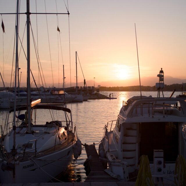 Marina al atardecer con una hilera de yates amarrados; mástiles, jarcias y banderas pequeñas se cruzan en líneas verticales contra el cielo cálido. Al fondo, el sol se posa sobre el horizonte detrás de una línea de montañas y la silueta de un faro. La luz dorada empata por unos minutos a los barcos caros con los baratos.