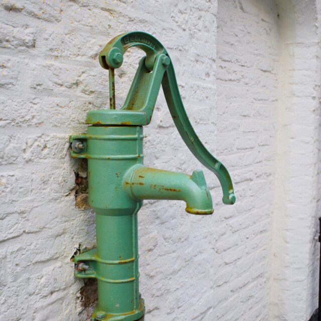 Bomba de agua manual antigua, pintada en verde mar y oxidada en las juntas, sujeta a una pared encalada con tornillos pasantes. La palanca curva descansa hacia abajo y un caño grueso espera el siguiente cubo. La pieza sigue ahí décadas después de que el grifo de la cocina haga el trabajo.