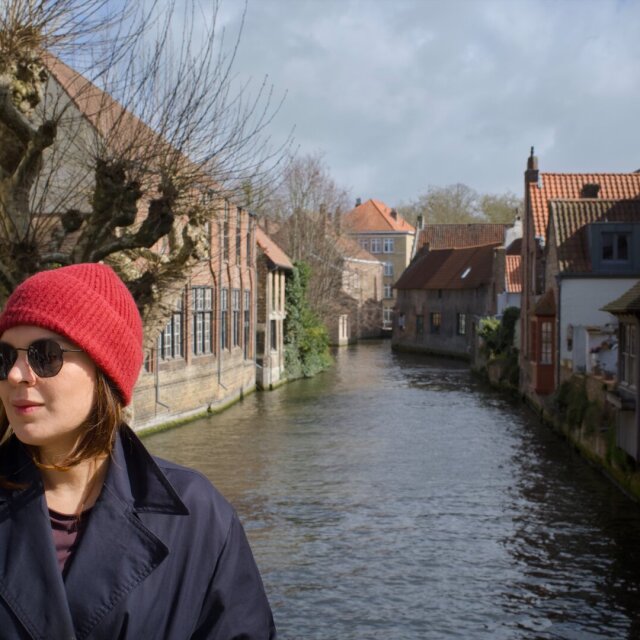Una mujer joven con gorro rojo de lana y gafas de sol mira hacia el lado, en perfil de tres cuartos; lleva una gabardina azul oscuro abierta sobre una camiseta morada. Detrás, un canal estrecho ordena la perspectiva entre casas de tejado a dos aguas y árboles podados en bola. El gorro concentra todo el calor del cuadro.