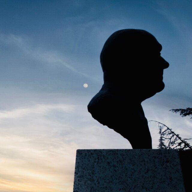 Busto de bronce sobre pedestal recortado en silueta contra el cielo del atardecer. La luna llena cuelga a la altura del rostro vacío, una rama de cedro entra por la derecha. Algunos retratos esperan a que les hablen, no al revés.