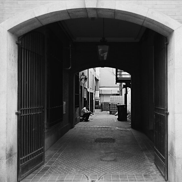 Pasaje con arco grande de piedra que se abre hacia un patio trasero; las dos hojas de la reja están abiertas y dejan ver al fondo una persona sentada en un escalón, con un libro o un papel en las manos. Junto a la pared, un contenedor de plástico. La calle vista desde dentro: el umbral mira hacia fuera y dentro hay alguien leyendo.