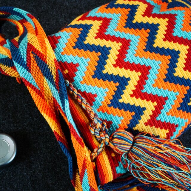 Bolsa de tejido tradicional con patrón en zigzag de colores saturados: rojo, naranja, amarillo, azul cobalto y turquesa se alternan en líneas dentadas que cubren toda la superficie. De la boca cuelga una borla larga de flecos del mismo cromatismo. A la izquierda, la tapa de un objetivo de cámara recuerda quién mira.