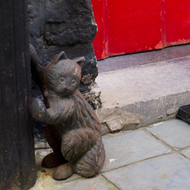 Un raspador de barro en forma de gato pequeño, fundido en hierro oscuro y oxidado, se apoya contra el quicio de piedra negra junto a un poste también de hierro. Detrás se asoma el panel rojo brillante de una puerta. El gato lleva décadas haciéndose el sorprendido sin que nadie le pase la mano por encima.