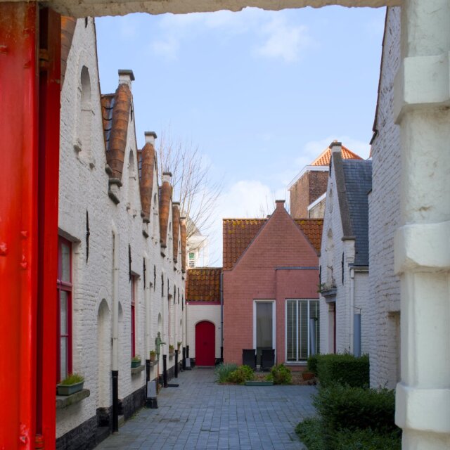 Vista a través de un marco rojo: un patio interior con casitas blancas de tejado escalonado se ordena a un lado del pasaje empedrado, y una pequeña vivienda de ladrillo rosa cierra el fondo con su puerta roja. La pintura del marco está descascarillada por años de portazos. El umbral hace de cuadro y la calle, de pintura.