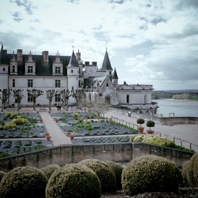 Un château de piedra clara con torres y tejados de pizarra se asienta sobre un parterre simétrico de grava, setos bajos y arbustos podados en bola. Detrás, un río ancho dibuja una curva con una isla larga. El cielo cargado le quita color a la mañana y deja la geometría del jardín como lo más decidido del cuadro.
