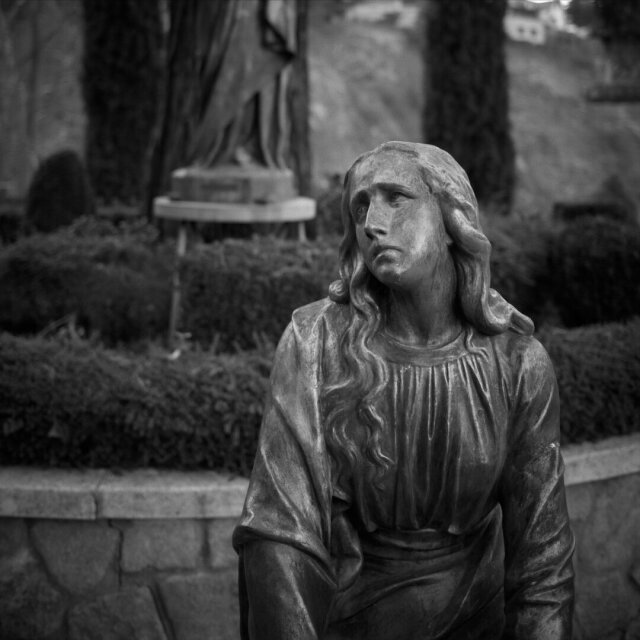 Una estatua funeraria de mujer con cabello largo y túnica lleva la mirada hacia un lado, los ojos vacíos por la erosión. Detrás, otra figura encapuchada se mantiene en pie sobre un pedestal junto a setos bajos podados en lecho rectangular. La piedra recoge cada arruga del paño que se hizo para no envejecer.