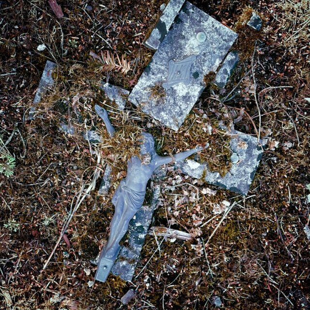 Un crucifijo metálico fragmentado yace sobre el suelo musgoso de un cementerio abandonado: la figura del Cristo sigue clavada en la pieza vertical mientras los brazos de la cruz están rotos a su lado. Hojas secas y pequeños líquenes han empezado a entrar por las grietas. La intemperie deshace los símbolos antes que las creencias.
