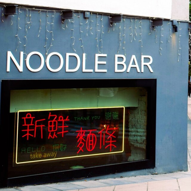 Fachada gris azulado de un Noodle Bar; en el escaparate, un neón rojo y verde superpone caracteres chinos a un saludo en inglés y otro en mandarín. Sobre el letrero, una hilera de luces de Navidad cuelga deshilachada en pleno día. La acera está enlosada, vacía a esa hora; al fondo se intuye una avenida arbolada.