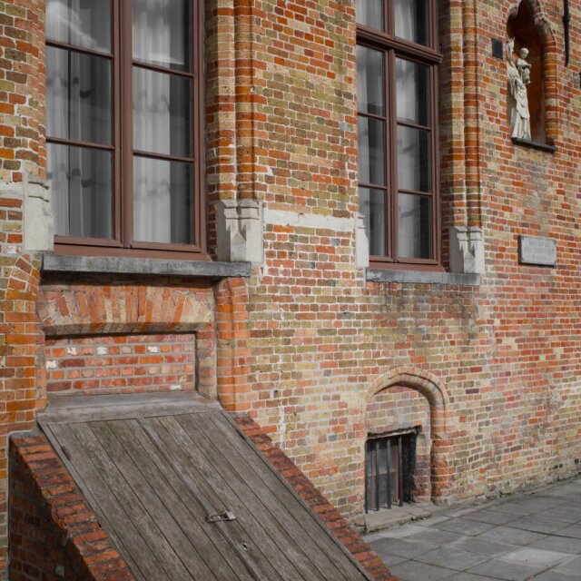 Fachada de ladrillo rojo gastado con dos ventanas altas, una hornacina con una pequeña Virgen y una rampa de madera inclinada que sale del muro como un cajón abierto. La piedra del zócalo se ha vuelto blanca por la humedad de los años. La rampa servía para subir mercancía a otra época.