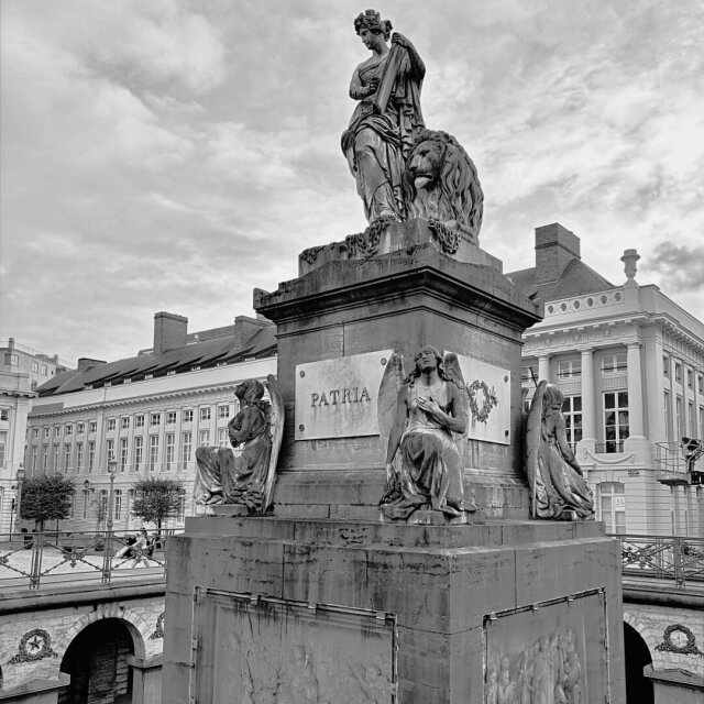 Monumento de piedra con figura femenina alegórica y león sobre el pedestal, en blanco y negro. Abajo, ángeles agachados flanquean una placa que dice PATRIA, edificios neoclásicos cierran el fondo. Las palabras grandes pesan más cuando las sostienen ángeles tristes.