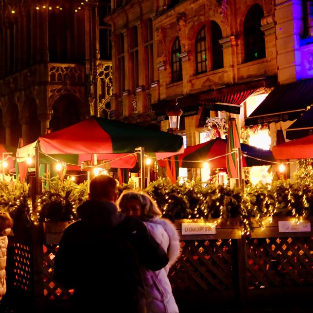 Mercadillo navideño nocturno: pareja abrazada en primer plano frente a casetas con toldos a rayas rojas y verdes iluminadas con guirnaldas. Detrás, una fachada gótica de oro viejo cierra la plaza. El frío se aguanta mejor cuando lo decoran.