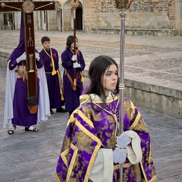Cofradía en procesión al atardecer: en primer plano, una joven con dalmática púrpura bordada en oro sostiene un asta plateada con farol. Detrás, tres nazarenos en hábito morado portan la cruz, uno con capirote blanco. La piedra de la plaza recoge el oro de la última hora.