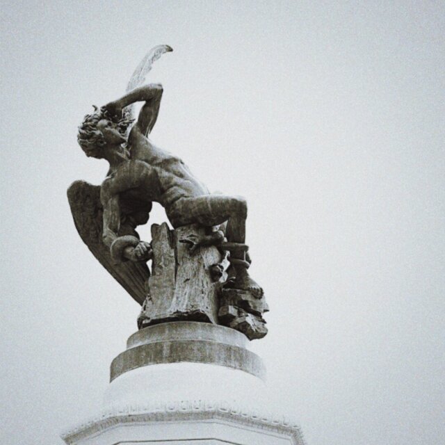Estatua del ángel caído sobre un tronco roto, una de sus alas extendida hacia abajo. La pose en escorzo retuerce el cuerpo como si aún estuviera cayendo, el cielo blanco lo deja sin profundidad. Hay esculturas que llevan un siglo sin terminar de tocar el suelo.