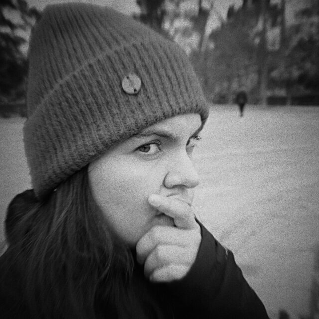 Retrato cercano en blanco y negro de una joven con gorro de lana de tejido grueso, dedo apoyado en los labios. Mira hacia un lado fuera del cuadro, pelo largo cayendo bajo la barbilla. La cámara también es una manera de pensar despacio.