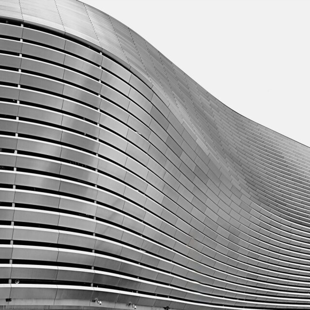 Fachada curva de un edificio moderno revestido de bandas horizontales de aluminio y cristal. Las líneas de las plantas dibujan una onda continua contra el cielo blanco, las sombras se acumulan en los pliegues. Algunas ciudades cambiaron de pulso el día que dejaron de pintar fachadas.