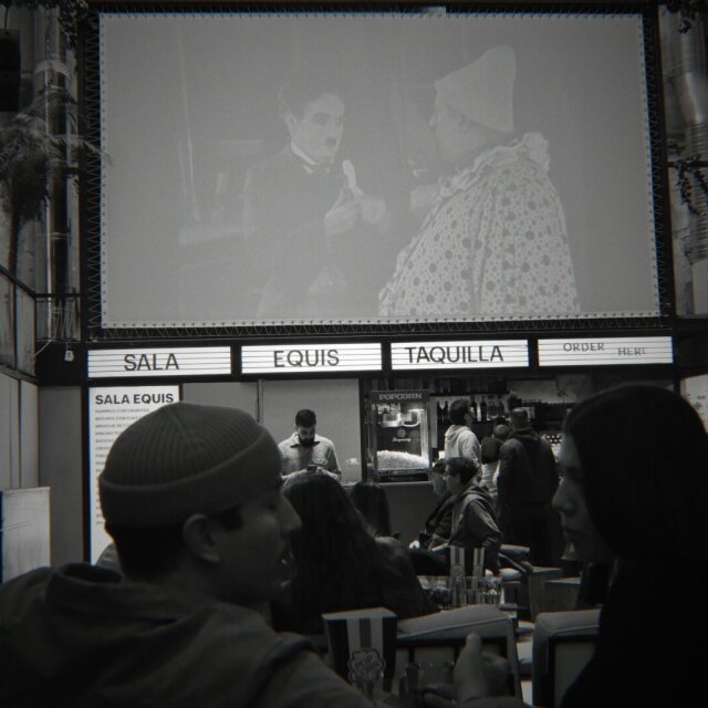 Interior de un cine-bar en blanco y negro: en lo alto, una proyección con Chaplin junto a un personaje encapotado en una escena del Klondike. Abajo, un rótulo retroiluminado anuncia SALA EQUIS, TAQUILLA, ORDER HERE, y dos cabezas en sombra ocupan el primer plano. Algunos lugares se llenan de gente porque la pantalla todavía las acompaña.