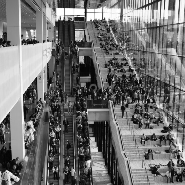 Interior en blanco y negro de un complejo de varios pisos con escaleras mecánicas cruzadas y gradas llenas de gente sentada con ordenadores. La fachada de cristal multiplica los reflejos hacia arriba, los cuerpos se vuelven hormigas en columnas. Hay multitudes que ya parecen una infografía.
