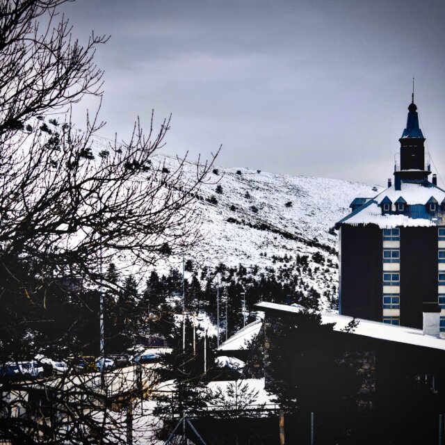 Edificio alto de tejado azul agudo sobre una ladera nevada, ramas peladas de un árbol cruzando el primer plano. El cielo plomizo iguala el blanco de la montaña, abajo se adivina un aparcamiento. La arquitectura de altura siempre está un poco a la defensiva.