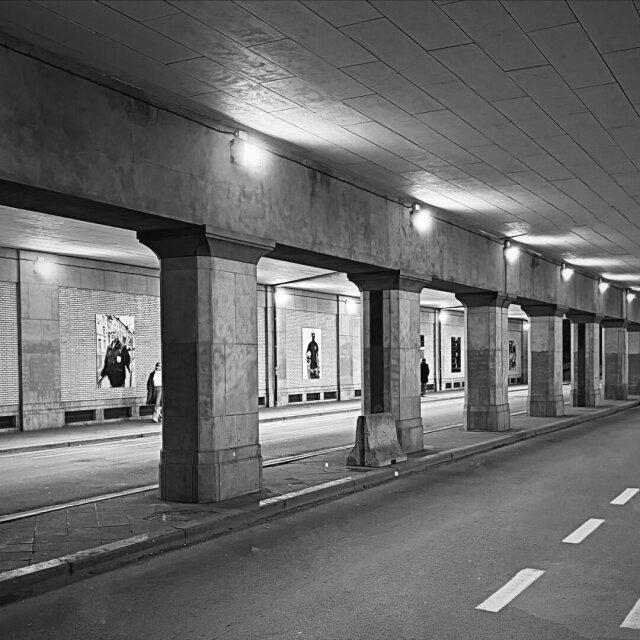 Pasaje cubierto en blanco y negro, columnas alineadas en perspectiva con cuadros de fotografía colgados en cada vano. La calzada vacía se extiende hacia el final iluminado, alguien lee una imagen al fondo. Las exposiciones públicas son los museos que sí madrugan.