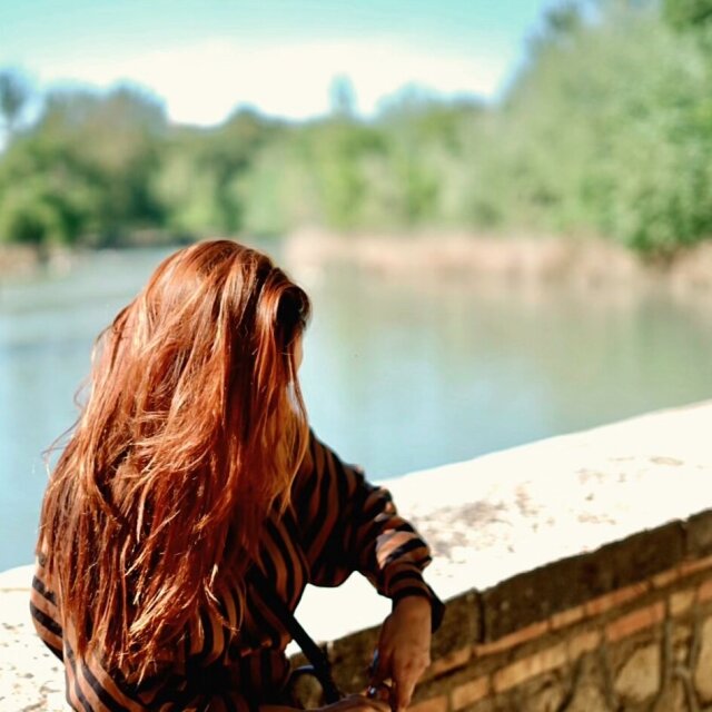 Mujer de espaldas, melena cobriza al sol, apoyada sobre un murete de piedra frente a un río de aguas verdes. Viste blusa de rayas marrones y negras, el follaje de la otra orilla está desenfocado. La luz del río se le sube al pelo antes que al pensamiento.
