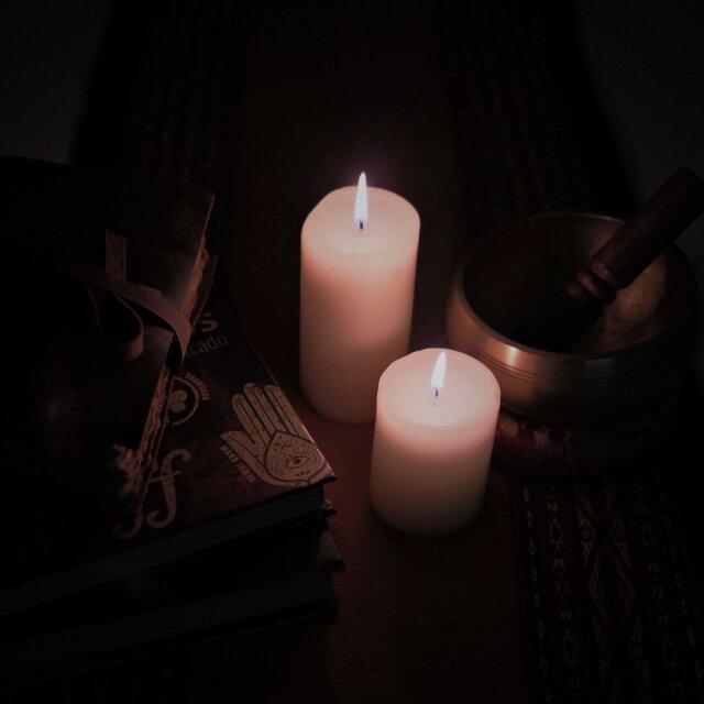 Dos velas blancas encendidas sobre una superficie oscura, junto a libros con una mano de Fátima en relieve y un cuenco tibetano de bronce. La penumbra come los bordes y deja sólo el contorno de la llama. Un altar improvisado puede ser cualquier mesa después de medianoche.