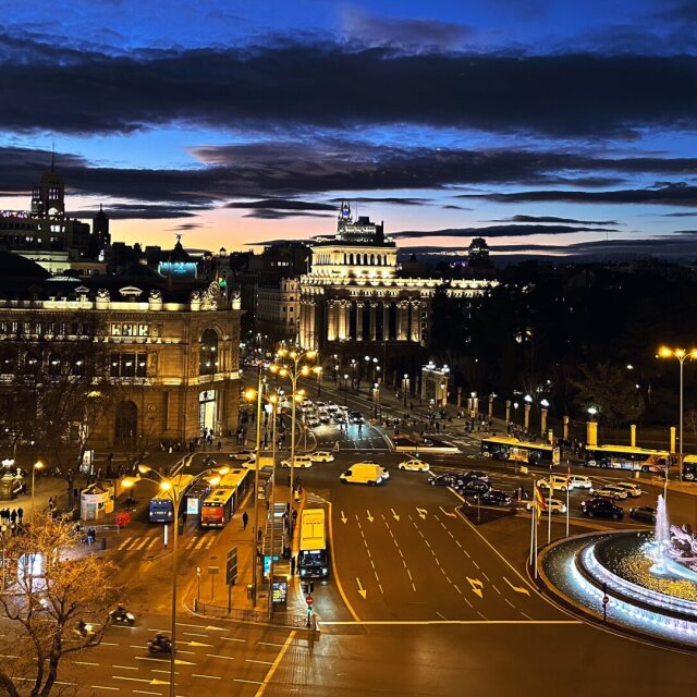 Plaza monumental al atardecer vista desde un balcón en altura, con fuente iluminada y una avenida de tráfico rodeándola. Las cúpulas y palacios al fondo recortan el último naranja del horizonte. Sólo la hora azul vuelve elegante hasta el atasco.