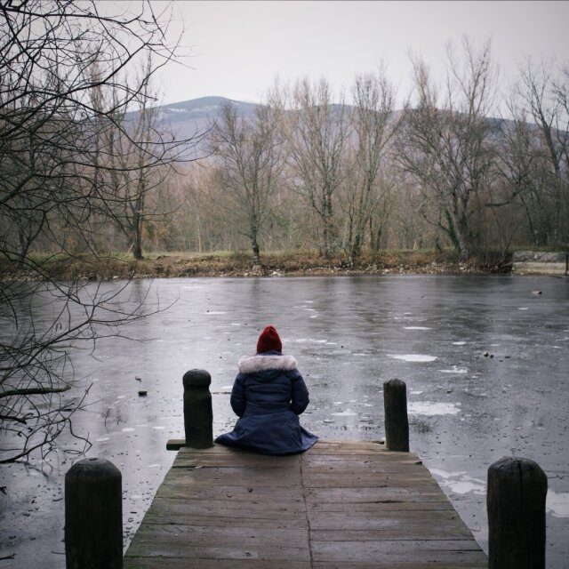 Persona de espaldas sentada al borde de un embarcadero de madera, frente a un estanque helado. Lleva parka azul con capucha de pelo y gorro rojo, las manos en los bolsillos. El silencio del hielo es de los que sólo se rompen mirando.