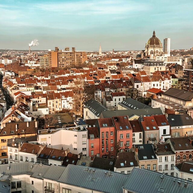 Vista en altura sobre tejados rojizos de una ciudad europea, una basílica con cúpula dorada despunta en el horizonte. Una chimenea industrial deja una columna de humo blanco a media distancia. Todos los barrios escogen un punto y todos lo señalan desde lejos.