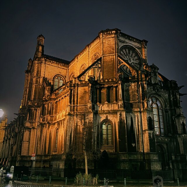 Catedral gótica vista desde su flanco, iluminada en tonos cobre contra el cielo casi negro. El rosetón, los contrafuertes y los pináculos se sostienen como una arquitectura de fuego frío. La piedra sólo deja de pesar por la noche.